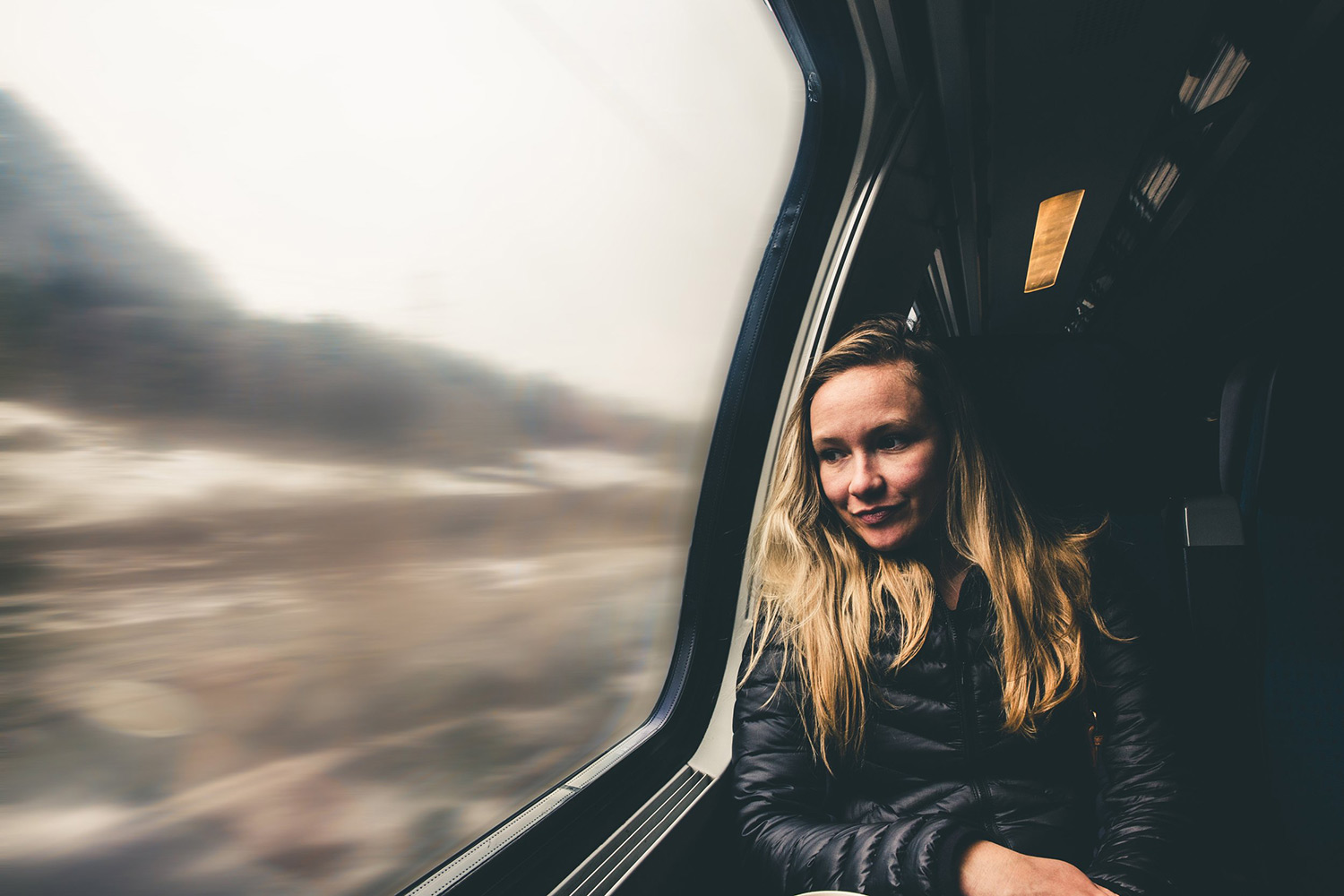 A blond woman on a train leans against the window, looking out