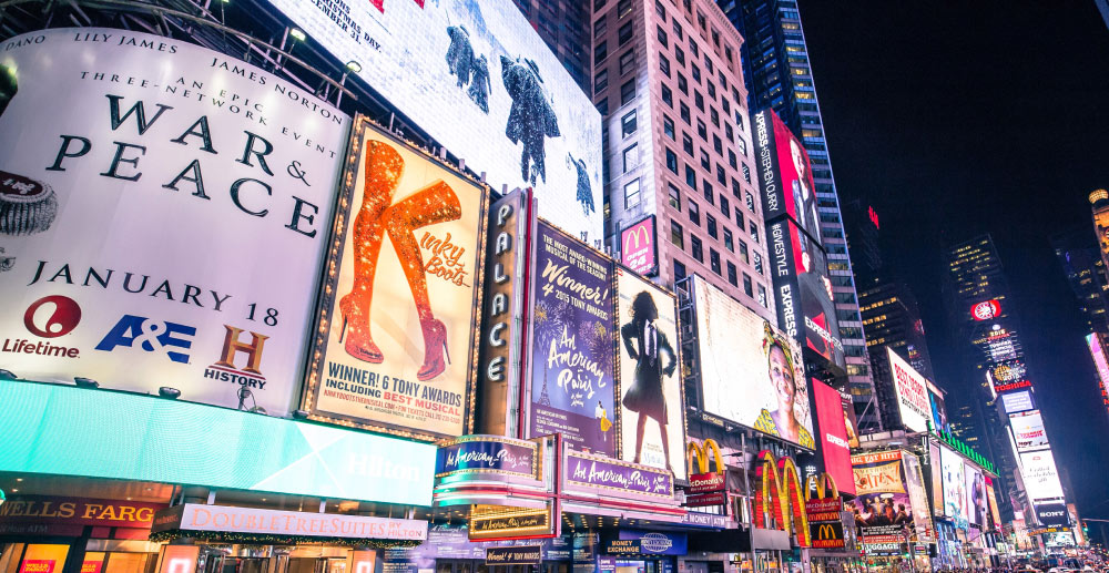 Broadway at night, lit up with signs for musicals and businesses.