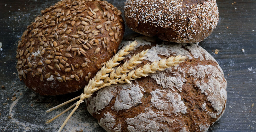 Three loaves of whole grain bread with a few stalks of wheat resting on top of them.