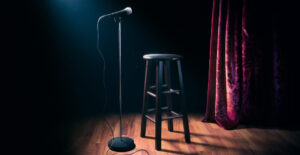 A black stool and microphone stand in a spotlight on an otherwise empty, dark stage.