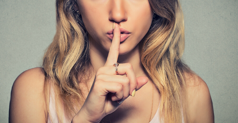 Young blonde woman holding a finger to her lips in a shushing gesture.