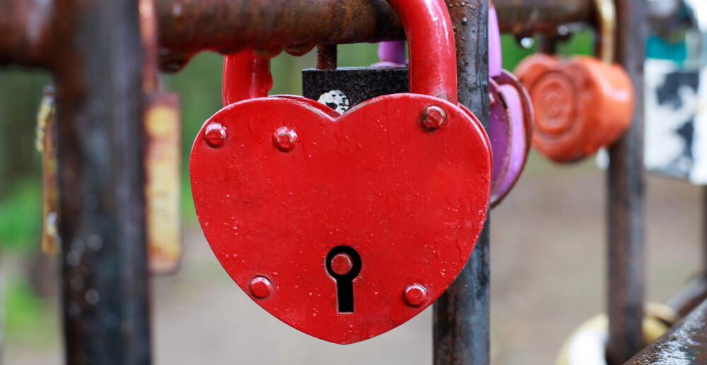A red, heart-shaped padlock locked to a railing that also holds other padlocks.