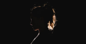 In a very dark space, a young woman is backlit so that her profile and ponytail are lit by a golden light, the only thing visible in the blackness.