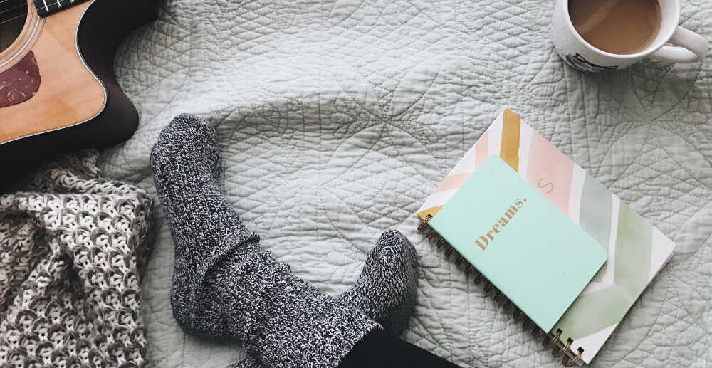 On a blanket-covered surface, a person's socked feet are crossed. An acoutsic guitar, dream journal, and mug of coffee sit nearby.