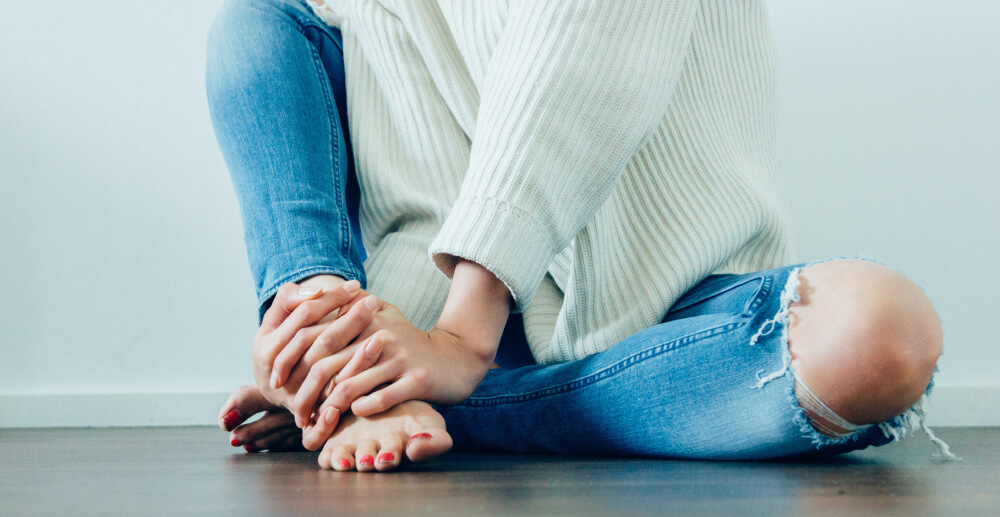 A woman in a white sweater and blue jeans with bare feet sits on the floor with one knee up, pulled close to her body. Her body is in frame but her face is not.