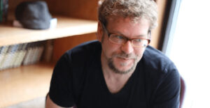 Author Daniel Maurer sits with a bookshelf behind him. He has curly hair and a goatee that are light brown. He wears glasses and a black t-shirt