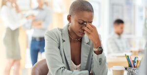 A young Black woman with close-cropped hair sits at a desk in a brightly lit office space. She pinches the bridge of her nose and looks distressed.