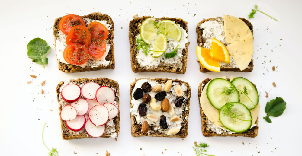 Six slices of whole whet bread arranged in a grid. Each is topped with goat cheese and additional ingredients, ranging from tomatoes to radishes to nuts.