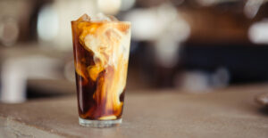 A glass tumbler of iced coffee with cream swirling through it.