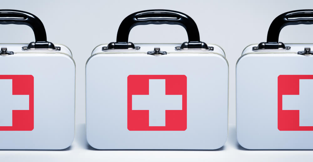 Three white, metal First Aid kits with red cross logos on the front of them. The background is a white counter against a white wall.