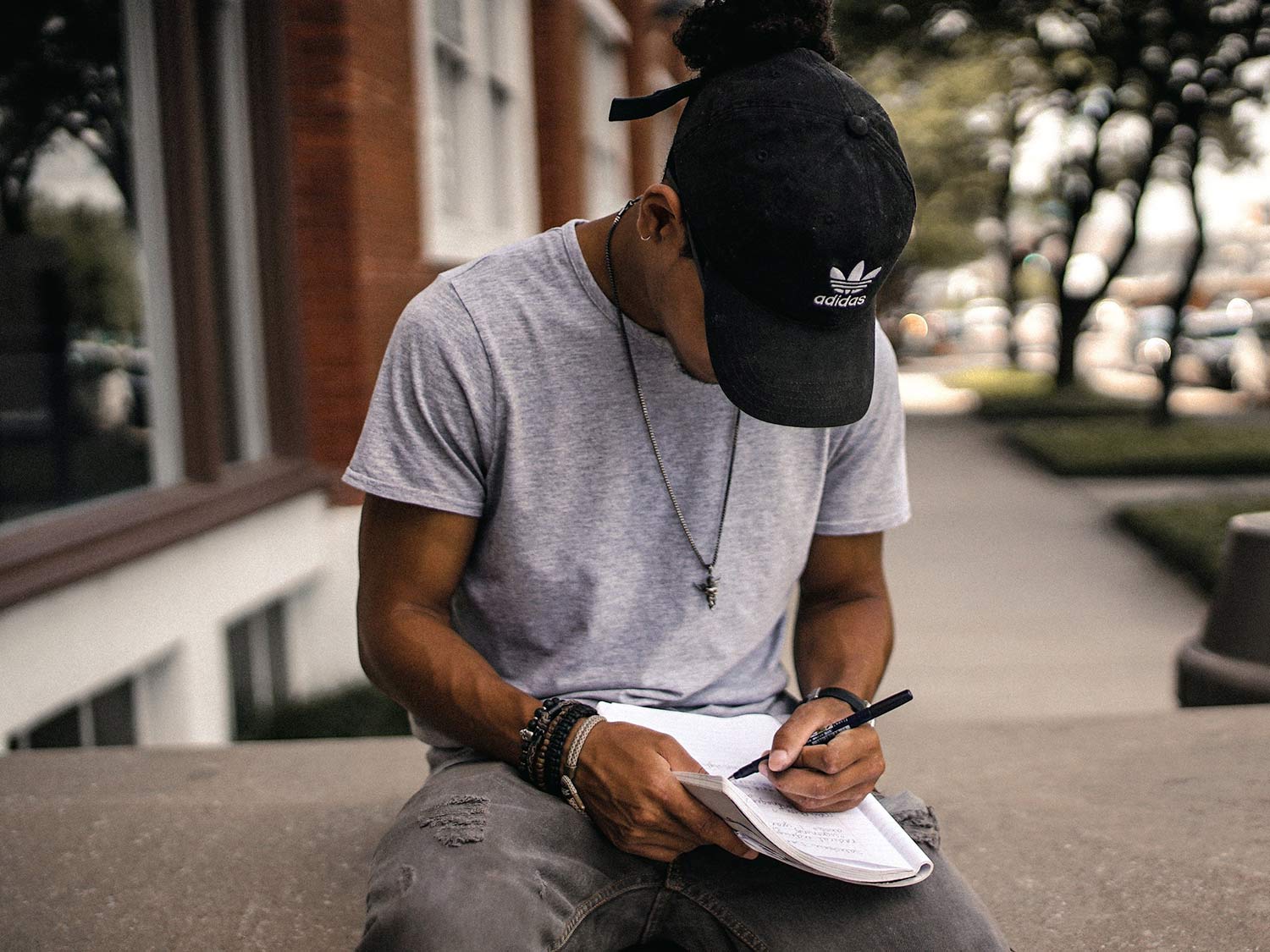 A man sits outdoors, writing in a notebook balanced on his leg