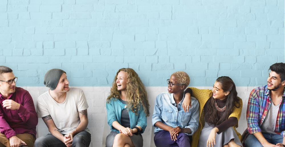 A row of people sit against a cinderblock wall, smiling and laughing together.