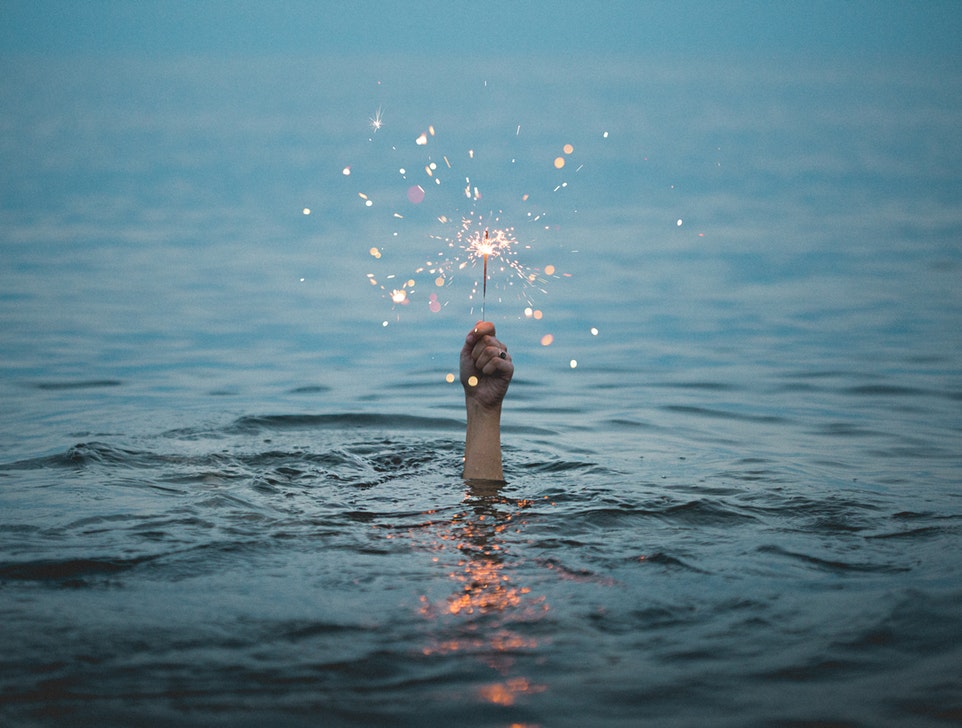 A hand reaches up out of the water, holding a lit sparkler. The rest of the body is submerged. Negative self talk.