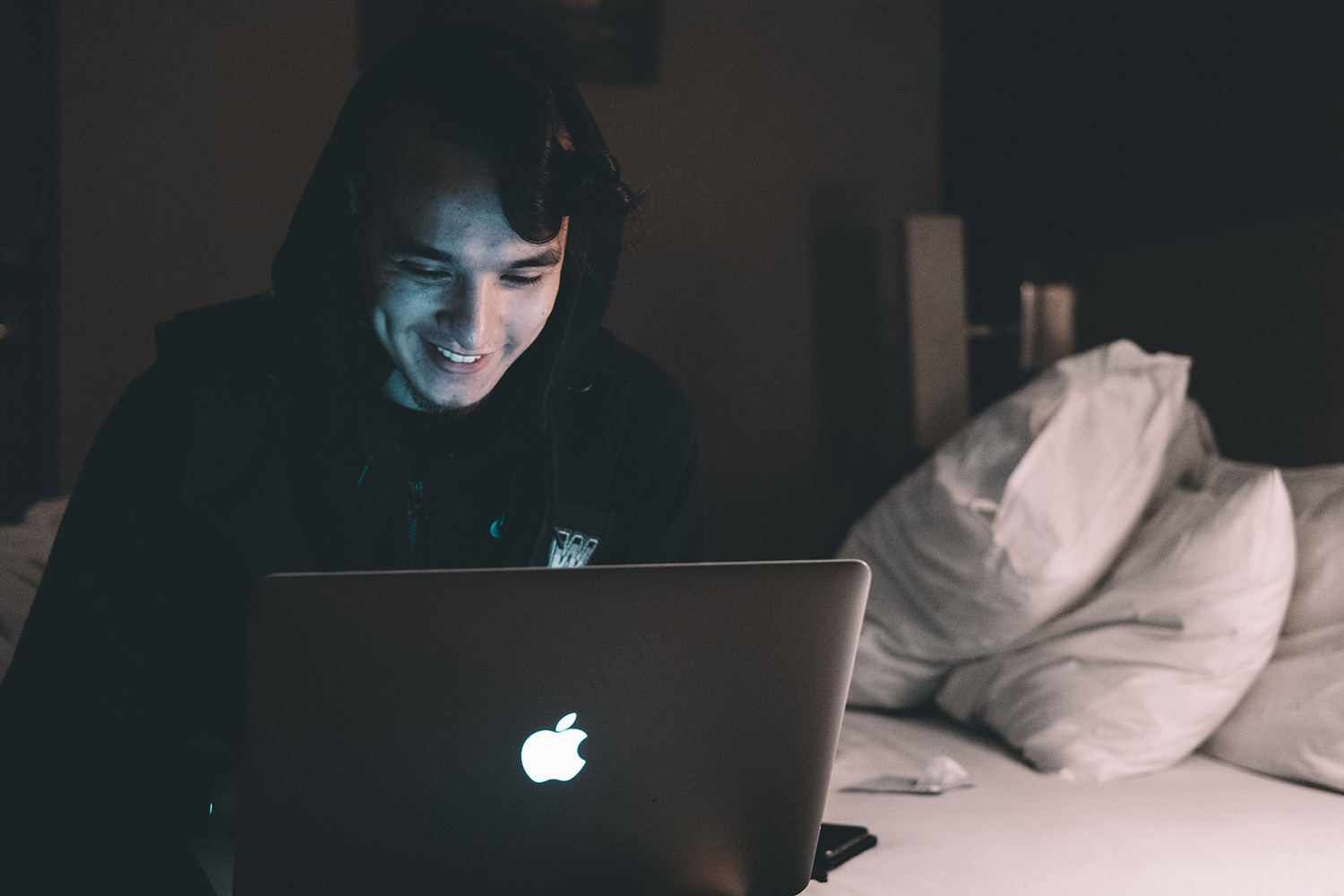 A smilking man uses a laptop while sitting on a bed. Online addiction treatment