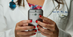 A female doctor in a white coat with a stethoscope around her neck uses a smartphone. The composition of the shot focuses on her hands, with her face out of frame.