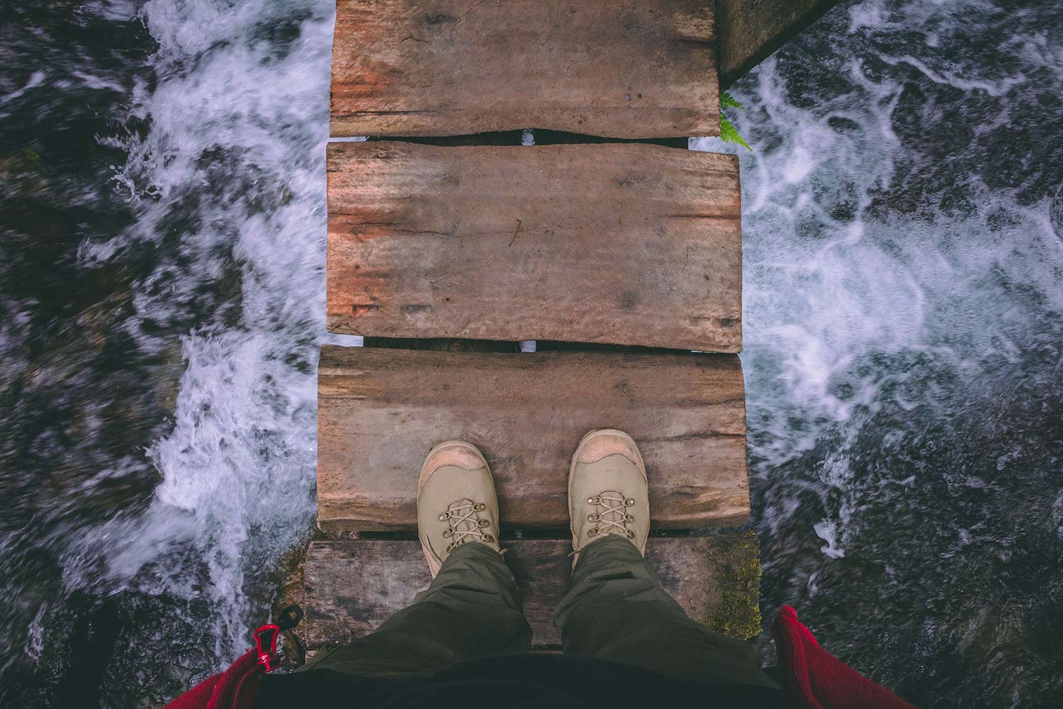 View down at a person's feet as they stand on a wooden plank bridge over a foaming river.