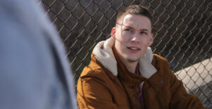 A young White man with short brown hair sits against a chainlink fence and looks up at a person who is out of focus and barely visible in the frame.