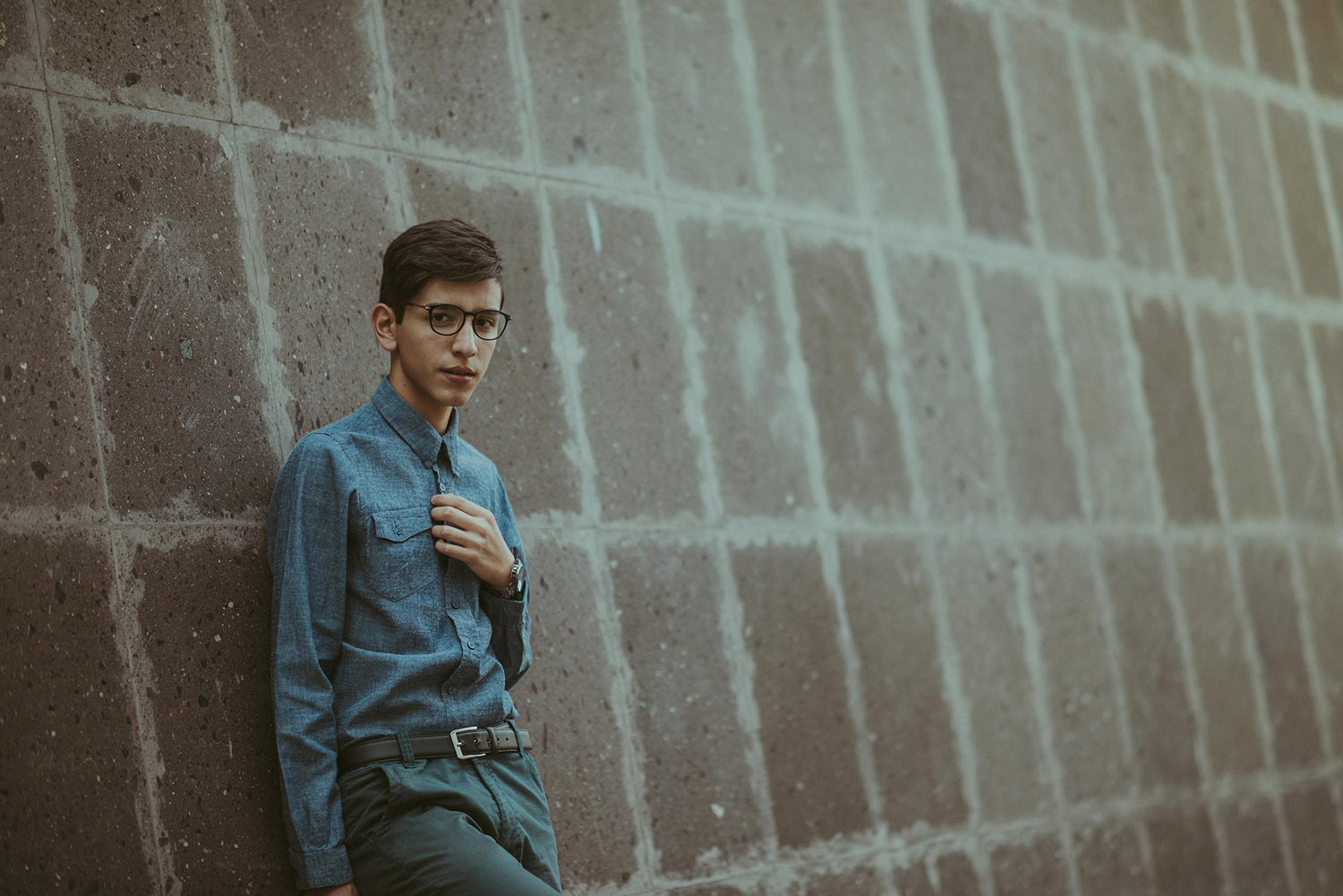 A teen boy in a blue button-up shirt leans against a concrete wall and stares warily at the camera.