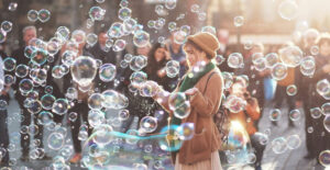 A woman wearing a knit hat, scarf, and jacket stands in a in a busy city space and laughs as she is surrounded by shimmering bubbles.