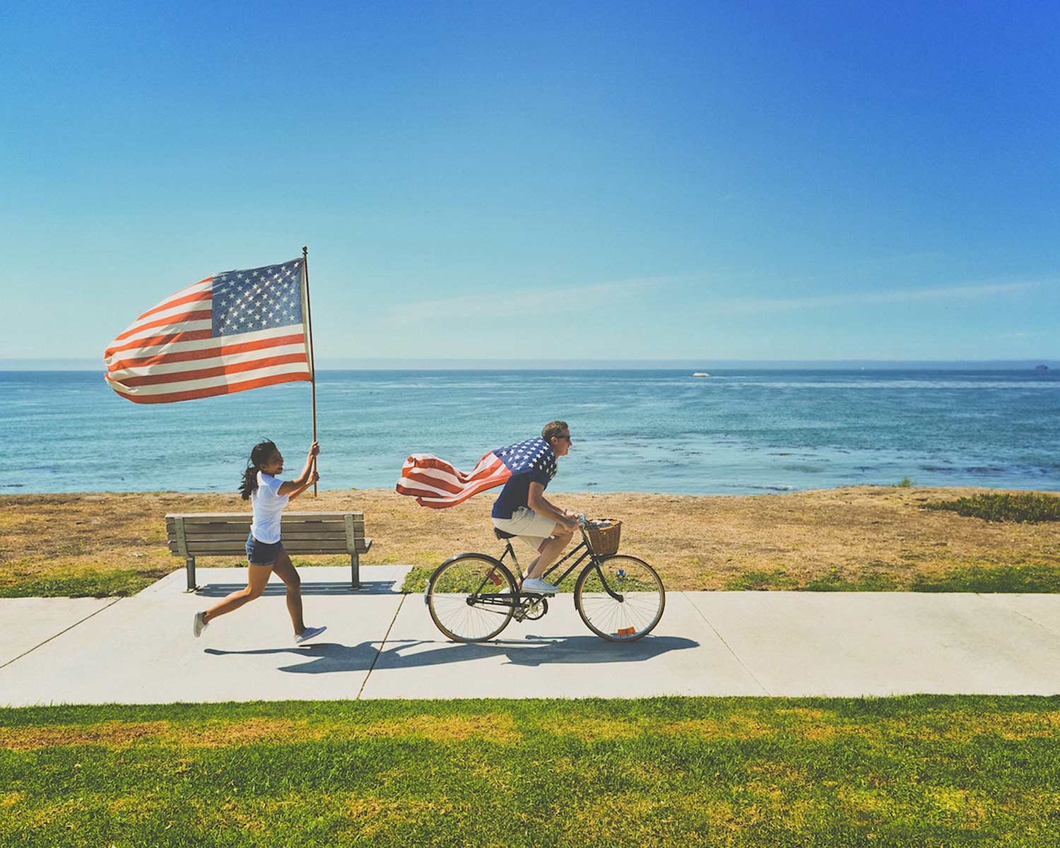 Beside the ocean, a man rides a bike with an American flag tied around his neck like a cape. Behind him, a woman runs with an American flag on a flagpole.