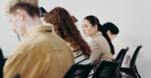 Office workers sitting in a row, seen from behind. One woman's face is in focus, as she looks toward her coworkers