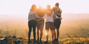 Four people with arms around one another stand atop a hill and look at the golden sunrise