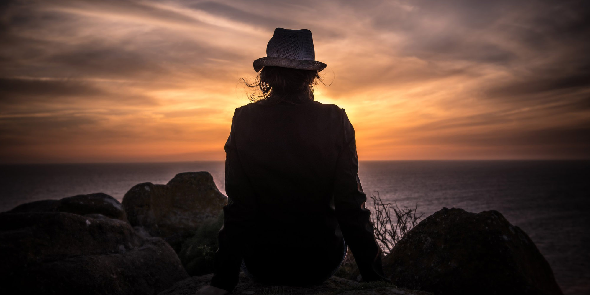 Rear view of a shadowed image. A woman in a hat stares out at the sunset.