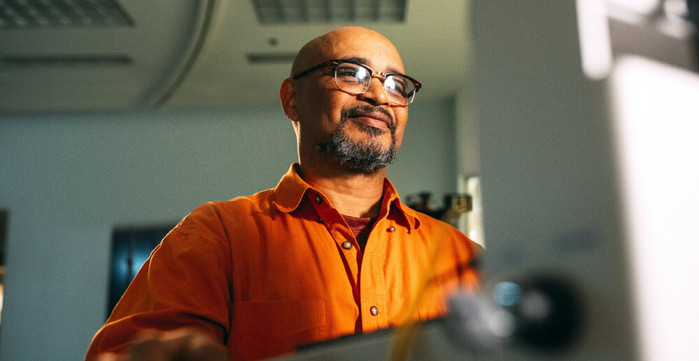 A man in an orange shirt and glasses stands at his work station facing a piece of electrical equipment.