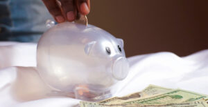 A clear plastic piggy bank sits on a white cloth. A brown-skinned hand is dropping a coin into the slot on the top of the bank, and there are $20 bills on the cloth in front of the bank.
