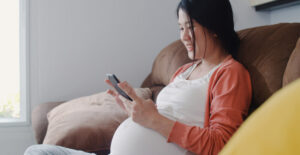 A young, East Asian woman who is clearly pregnant sits on a sofa and scrolls on her smartphone.
