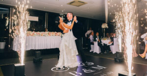 A bride and groom laugh as they dance alone at their wedding. Guests watch from their reception tables, and sparklers burn near the edge of the dance floor.