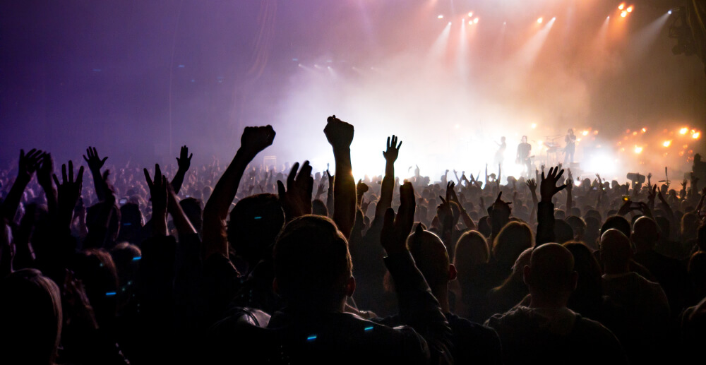 Rear view of the crowd at a concert with their hands in the air. The band is onstage in the distance, and the sky is hazy, with lights beaming through it.