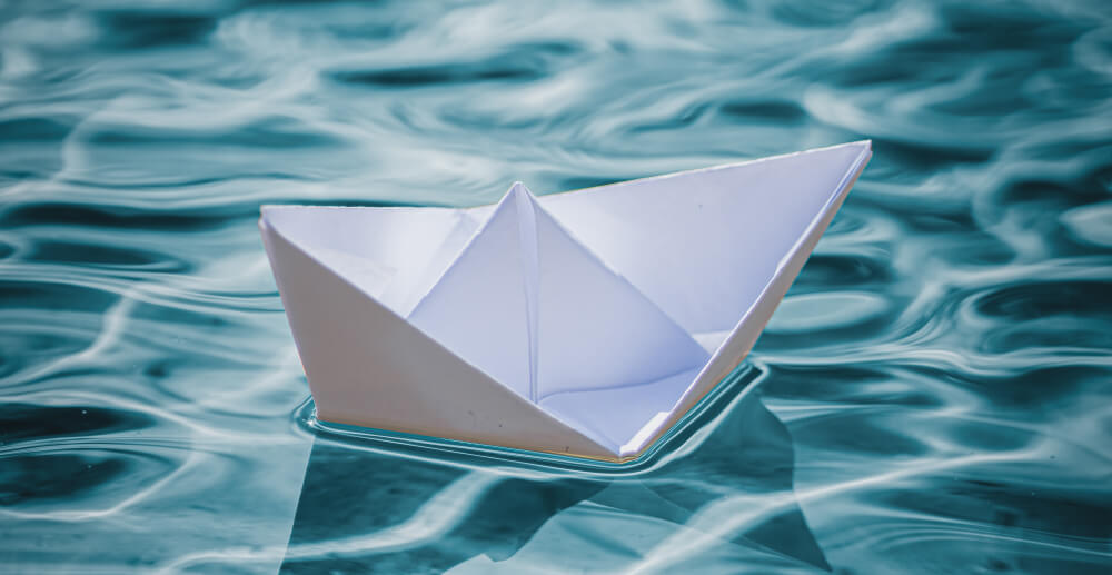 An origami boat folded from white paper floats in blue water with soft ripples.