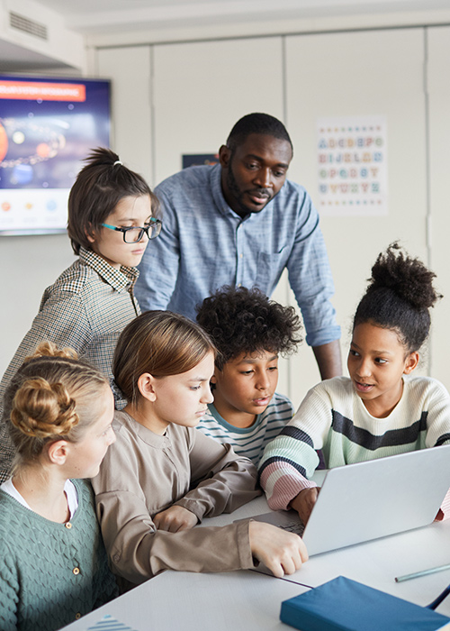 Male teacher supervising kids who are using a laptop