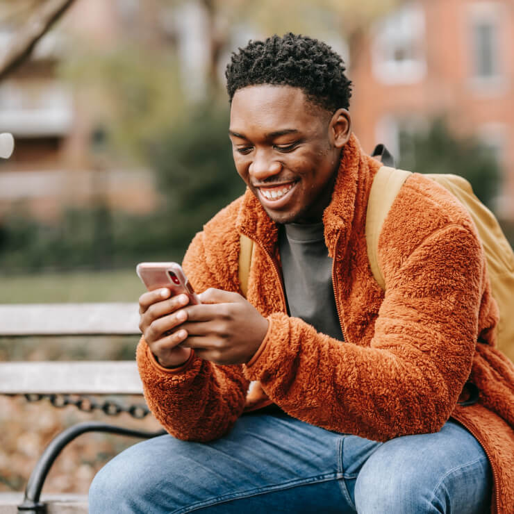 A young man wearing a fuzzy orange jacket and a backpack sits on a park bench as he smiles at the smartphone he's using.