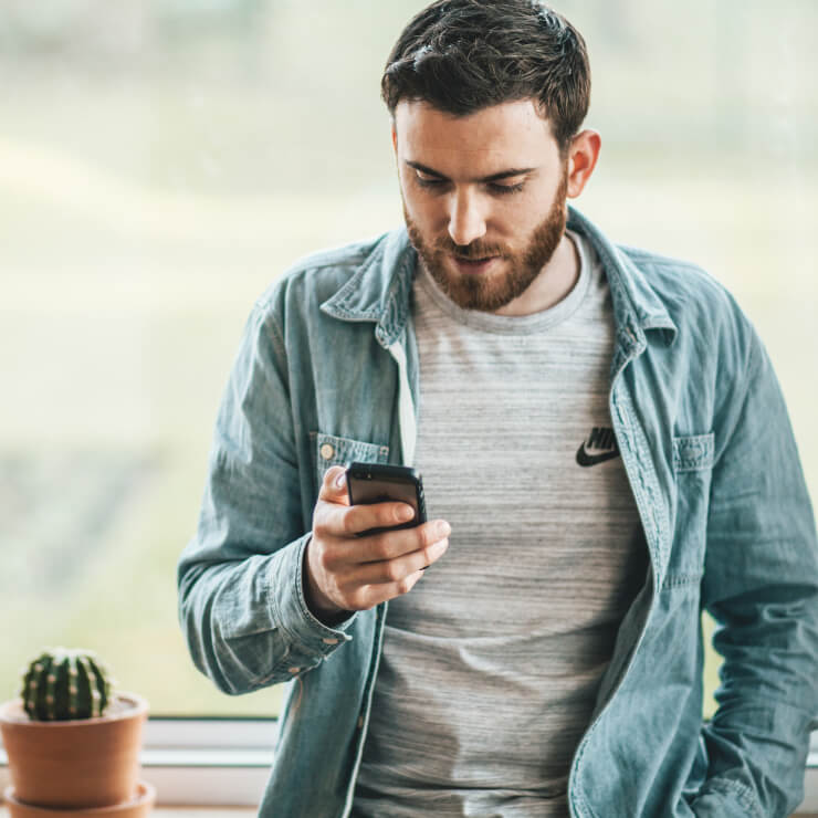 A young man with brown hair and a trimmed beard leans against a windowsill that holds a potted cactus. He is looking down at the smartphone he holds in one hand.