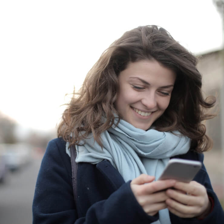 A young woman with brown hair and a blue scarf around her neck stands on the street in a small town, smiling down at her smart phone.