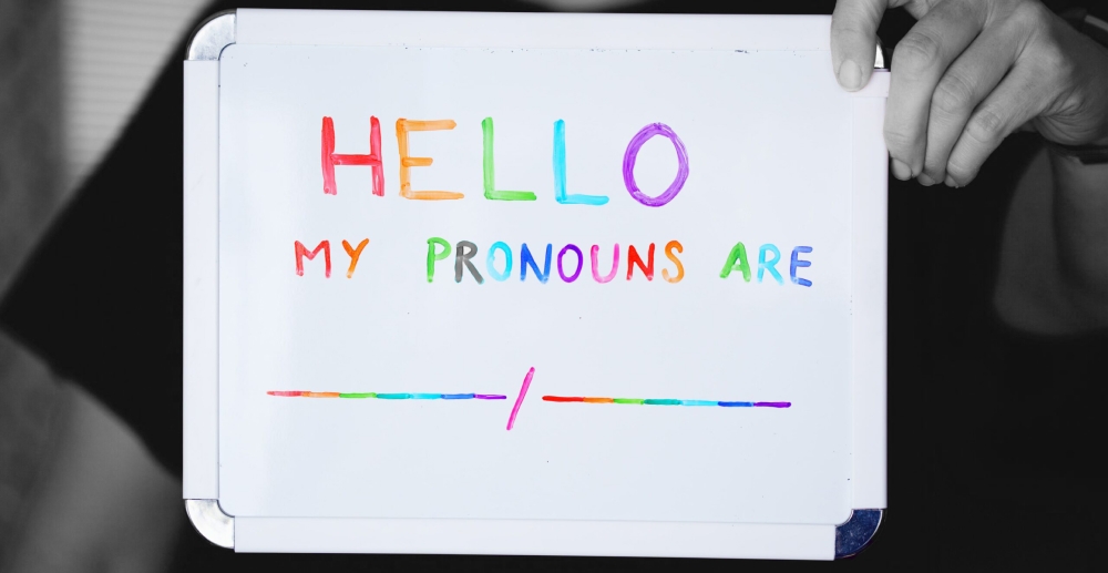 A person holds a small, dry-erase board with text written in rainbow markers "Hello, my pronouns are _/_". A Journey to Self-Love