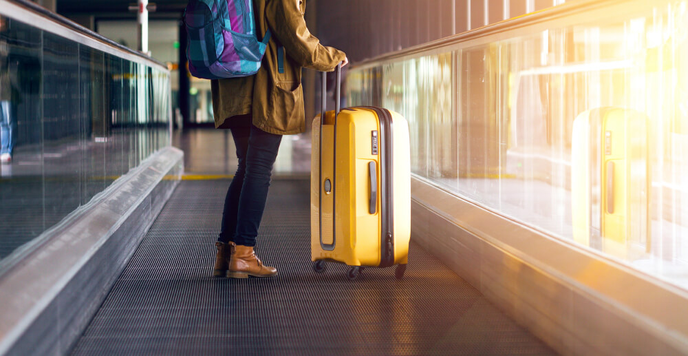 A woman with a yellow rolling suitcase and a blue and purple backpack stands on the moving walkway in an airport. Vacation sober, traveling sober.