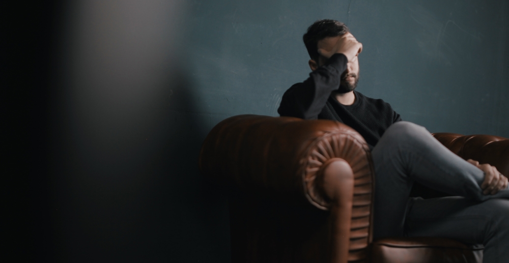 Darkly lit photo of a man sitting on a sofa holding his head in one hand in a posture of emotional distress.