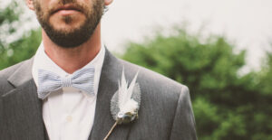 Closeup of a groom on his wedding day, wearing a gray suit and a white tie. He has a boutonierre made of feathers.
