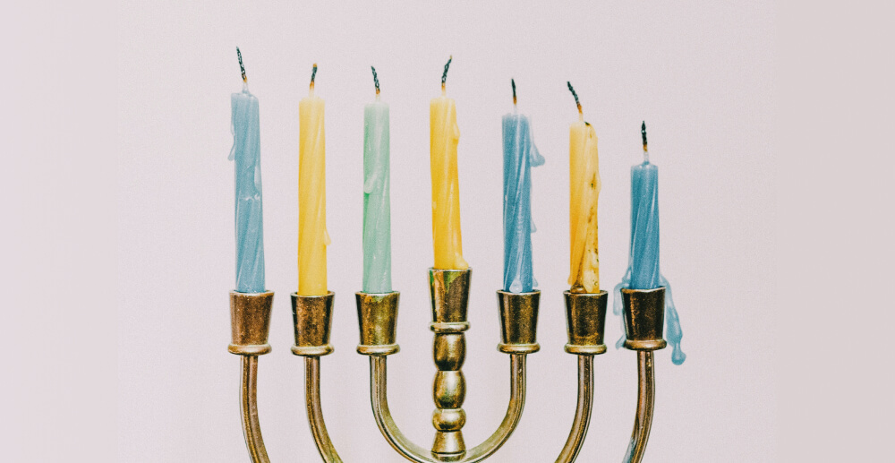 A brass menorah with seven blue and yellow candles. The candles are unlit but have darkened wicks and melted wax to show that they've been lit before.