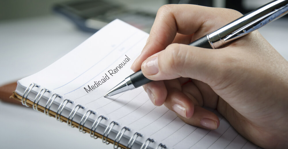 Closeup of a notebook, with a hand holding a pen to the page. The words, "Medicaid Renewal" are written.