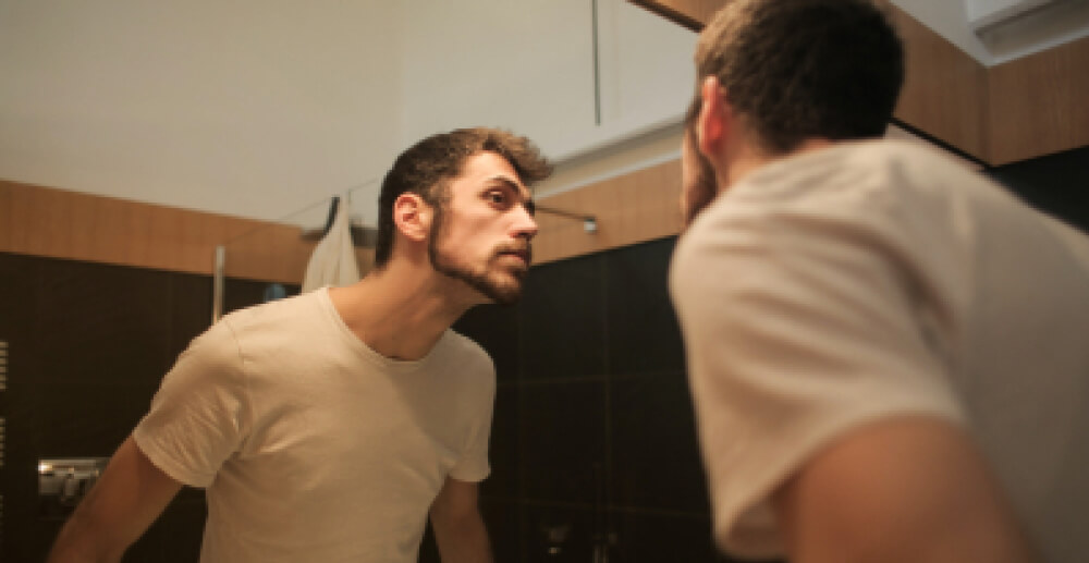 A young man in a white t-shirt looks at himself in the mirror