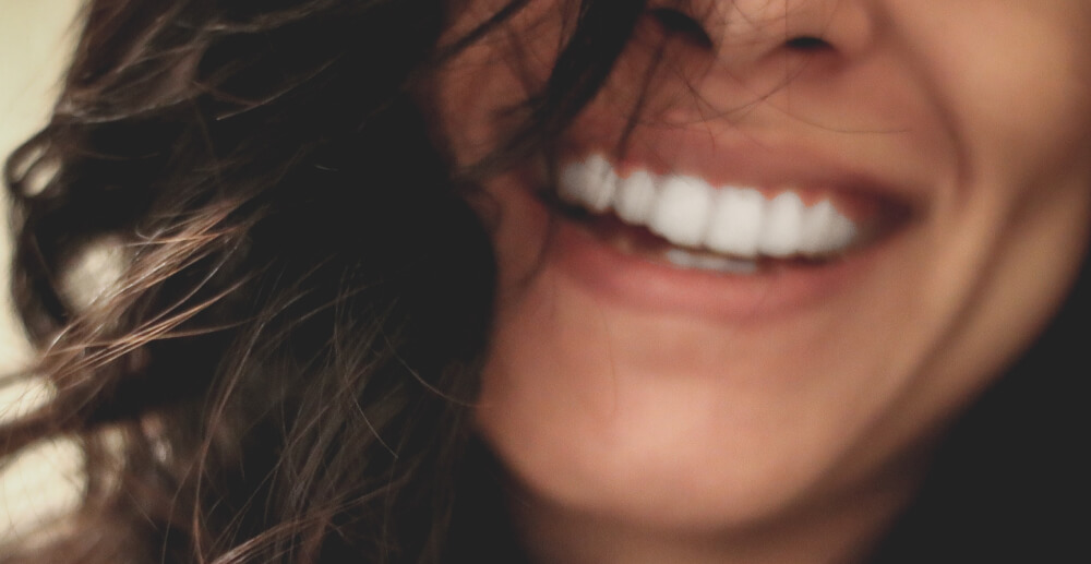 Close-up of a woman's smiling mouth. Her teeth are straight and white, and her long brown hair falls loose around her face.