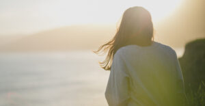 The back of a woman wearing a gray sweatshirt, sitting on a rocky shore and looking out at the sea. The sun is low and golden, as though it is very early morning or nearly sunset.