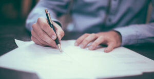 Closeup on the hands of a man in a long-sleeved, blue, button-up shirt as he writes on some papers with a ballpoint pen.