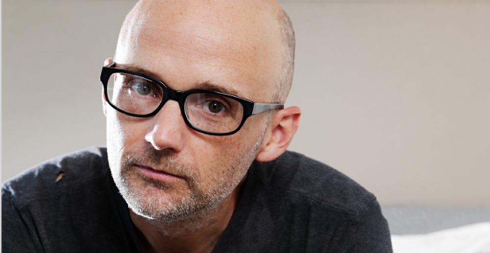 Portrsit of the musician Moby, who is a thin, white man with a shaved head and glasses. He has stubble on his face and wears a black t-shirt with a small hole on one shoulder.