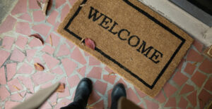 A welcome mat in front of the door to a house. The point of view looks down at the viewer's feet in front of the welcome mat.