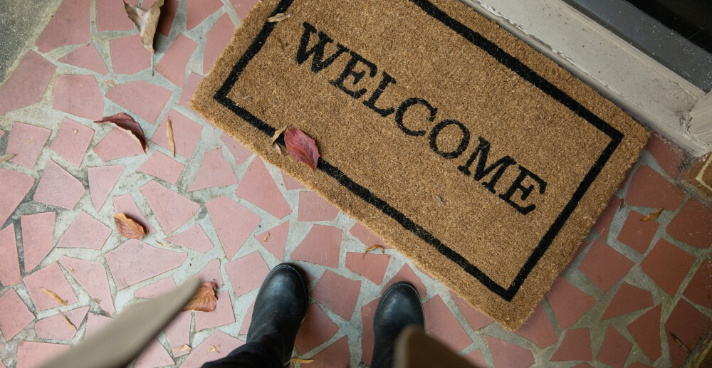 A welcome mat in front of the door to a house. The point of view looks down at the viewer's feet in front of the welcome mat.